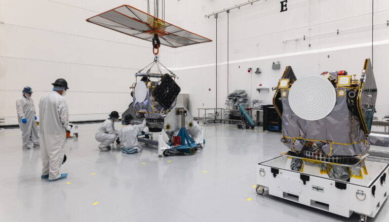 men in white suits in a white room working on an object suspended from a crane