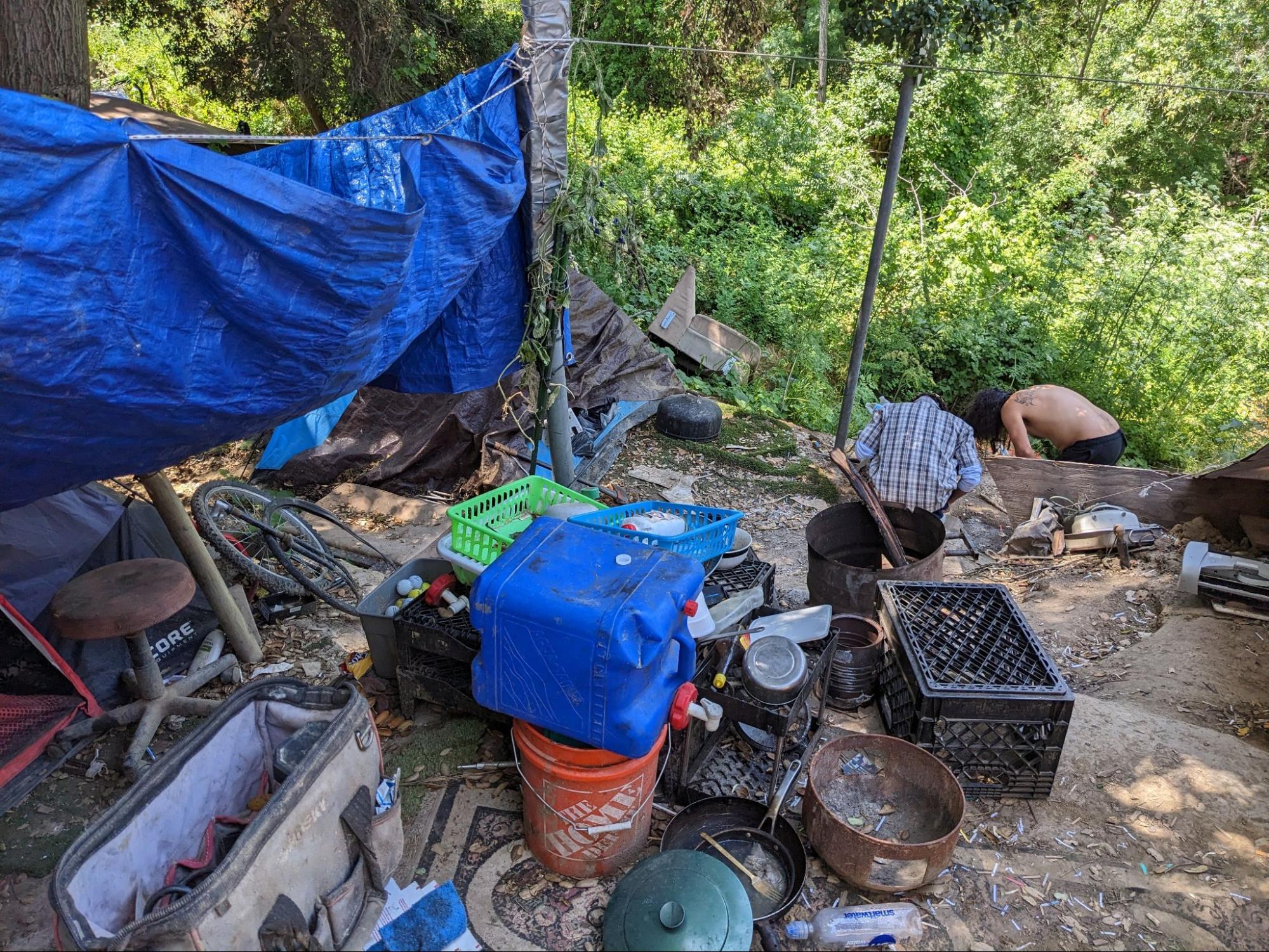 Two people look toward green bushes at a homeless encampment covered by a blue tarp, with items scattered on the ground