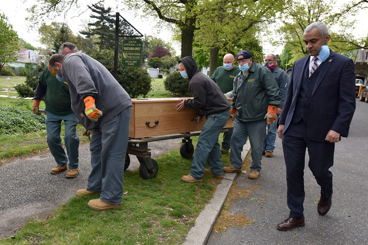 a man in a suit walks along with a wooden casket being rolled by workers in masks 