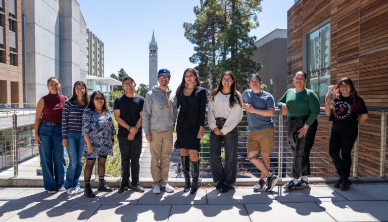 Nine students stand outside on the UC Berkeley campus, with the campanile visible in the background.