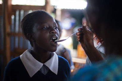 a woman pours medicine into a young African woman's mouth using a spoon