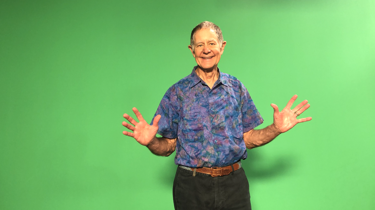 A person in a blueish shirt and dark pants standing with his arms extended in front of a green screen