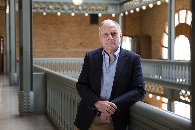 Nobel Prize winner David Card poses on the upper floor of the Hearst Mining Building, looking straight at the camera with his arm on the railing and his left hand clasping his right wrist.