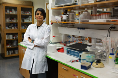 A photo of Kiana Aran leaning against a lab bench, wearing a white lab coat