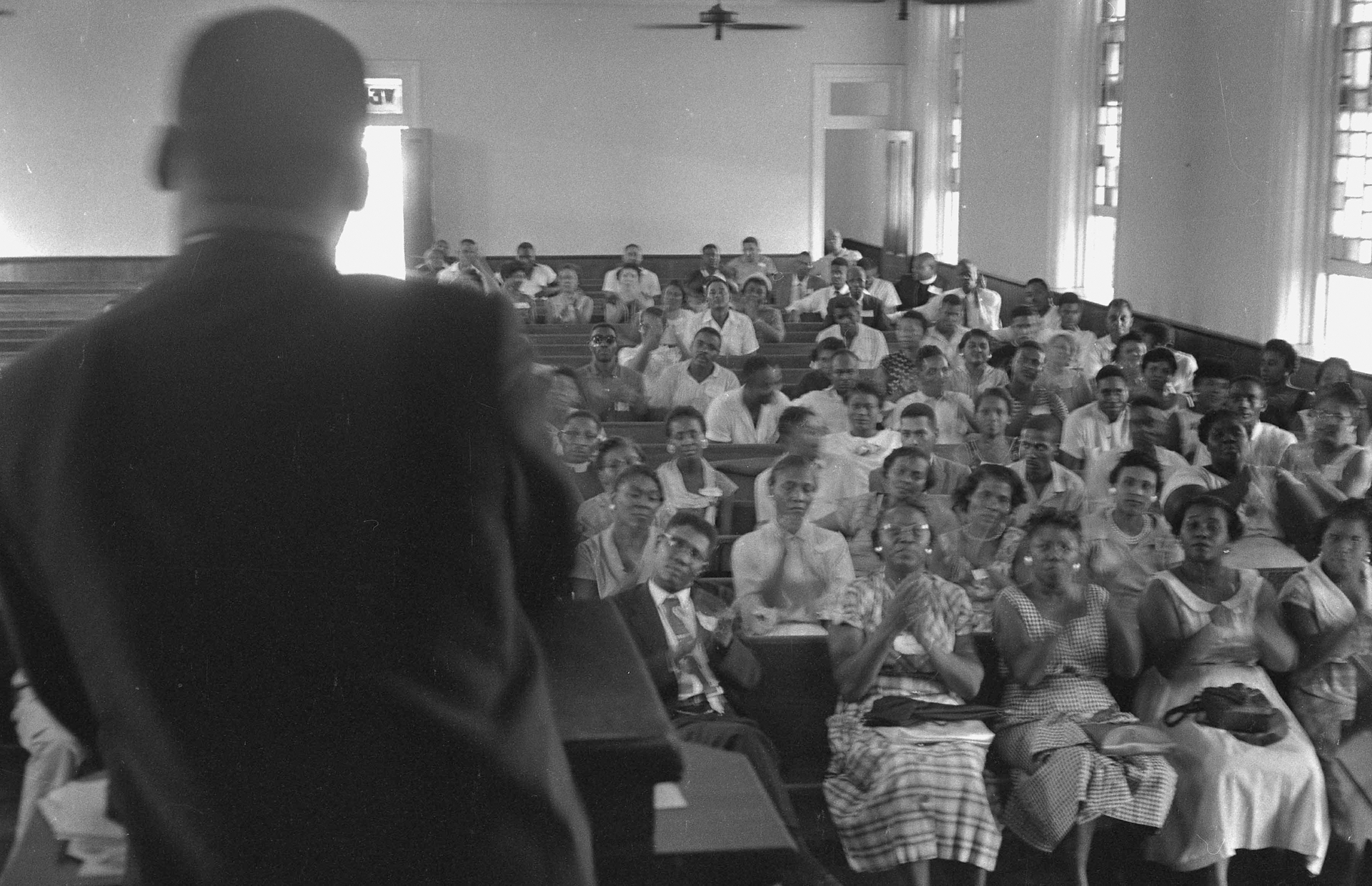 Black and white image of Martin Luther King Jr. with his back to the camera, and standing at a church pulpit speaking to a congregation of churchgoers sitting on church pews.