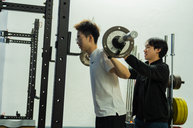 Christian Chhom assists a student with a barbell on his shoulders preparing to do a squat