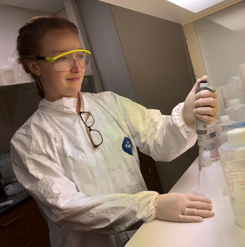Elizabeth Niespolo in white lab coat and goggles pipetting under a fume hood