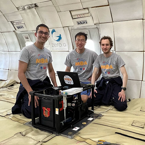 A photo shows three people in grey shirts sitting around an experimental box that has been secured to the floor of an aircraft cabin.