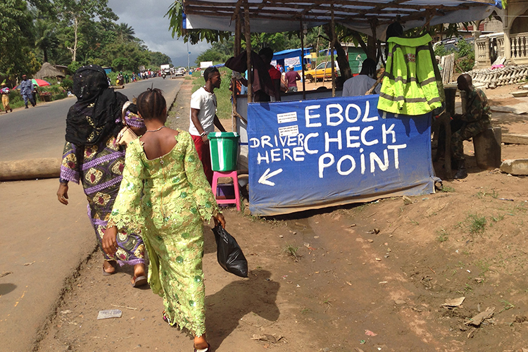 Women in the African nation of Sierra Leone walk past a streetside Ebola checkpoint