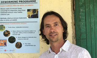 UC Berkeley economist Edward Miguel stands outside a building in Africa, next to a poster about a de-worming project