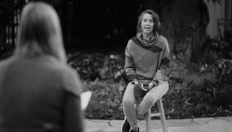 black and white photo of a woman seated on a stool talking to a woman in foreground, seen from behind