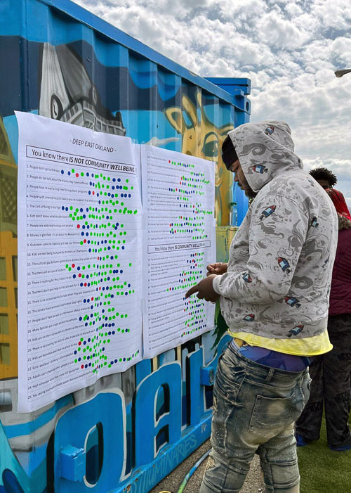 A young Oakland man at an outdoor event looks over a large poster that lists positive and negative indicators for community well-being  