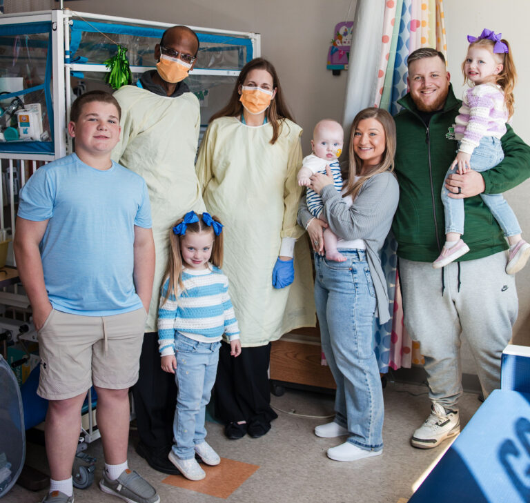 two doctors with masks posing with a family—mom, holding a baby; dad, holding a girl; a boy and a young girl.
