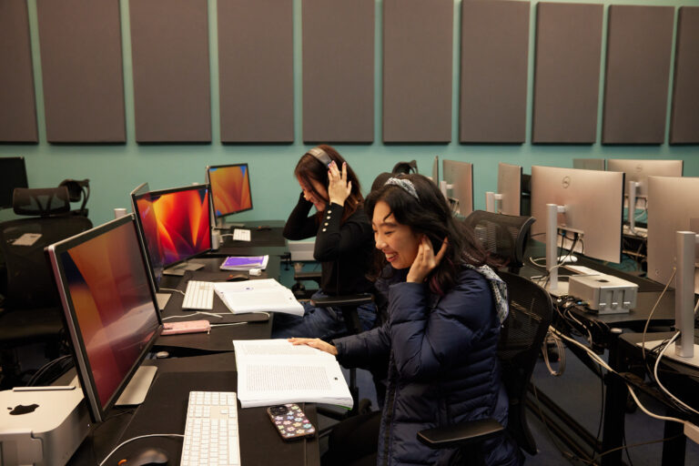 Two students study, in a campus media lab. They're wearing headphones and sitting at a table with microphones and computers.