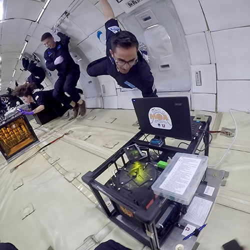 A photo shows an experimental box secured to the floor of an aircraft cabin while a person in a blue jumpsuit floats above it.