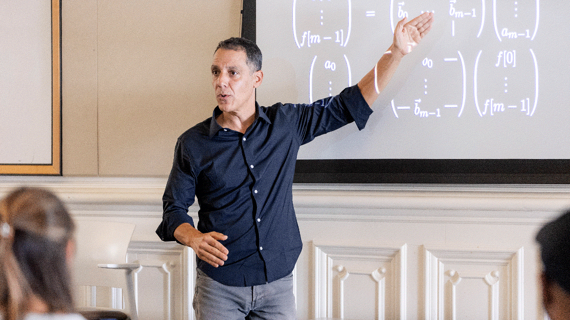 UC Berkeley professor Hany Farid stands at the front of a classroom, gesturing at equations projected onto a large screen 