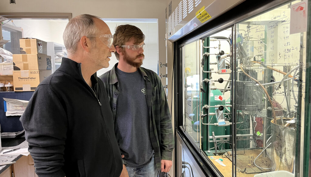 two men wearing goggles looking at an experiment behind the glass of a fume hood