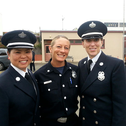 Three smiling women in uniform stand side-by-side on a city street.