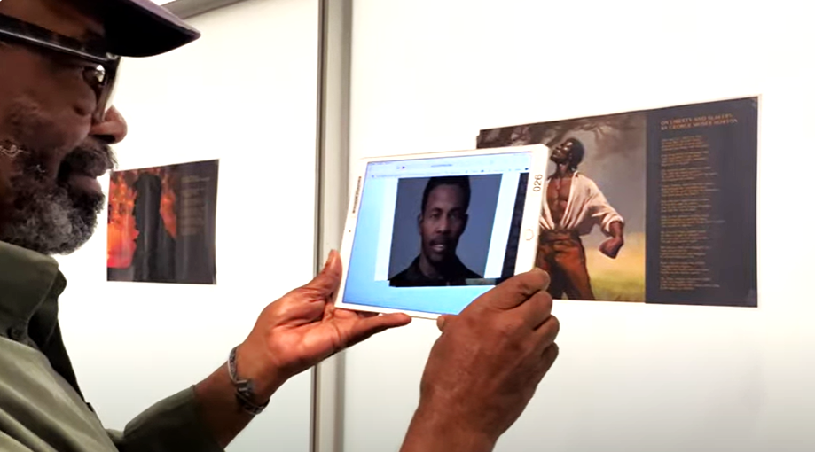 Cecil Brown holds up an iPad to an illustration of a Black slave displayed on a wall while a generative AI image of George Moses Horton displays on the digital screen.