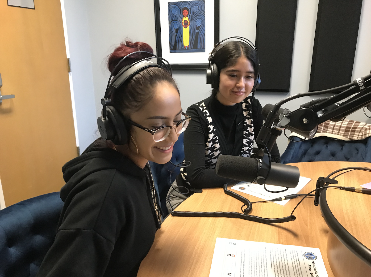 Two students smile as they speak into a microphone while sitting at a table. They are also wearing headphones and speaking as part of a podcast.