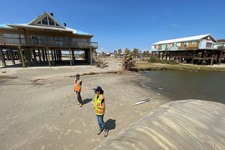 A photo of two people wearing safety vests, standing on a sandy beach next to a house on stilts