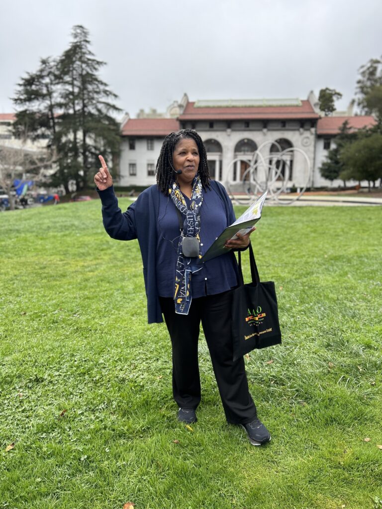 Wearing a Berkeley scarf and a blue shirt, Gia White holds up a finger as she talks. She's standing on the grass in front of an impressive campus building and holding a binder of information in her other hand.