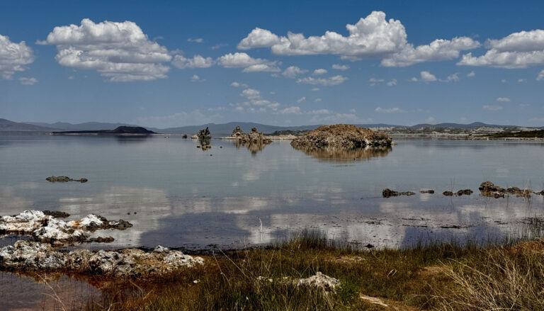 still water reflects blue sky and white clouds, with small islands in the lake and white crusty mounds in foreground