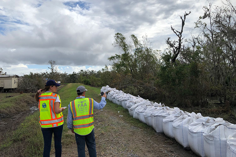 Two people wearing safety vests stand in a field next to a pile of sandbags.