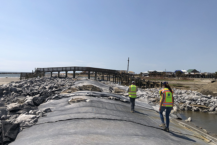 A photo shows two people wearing bright safety vests walking along a long, low levee by the coast on a bright clear day.