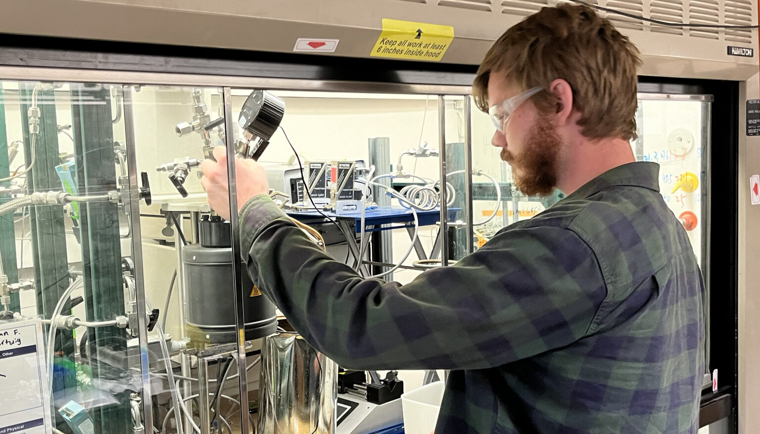 a bearded man wearing goggles adjust equipment inside a fume hood