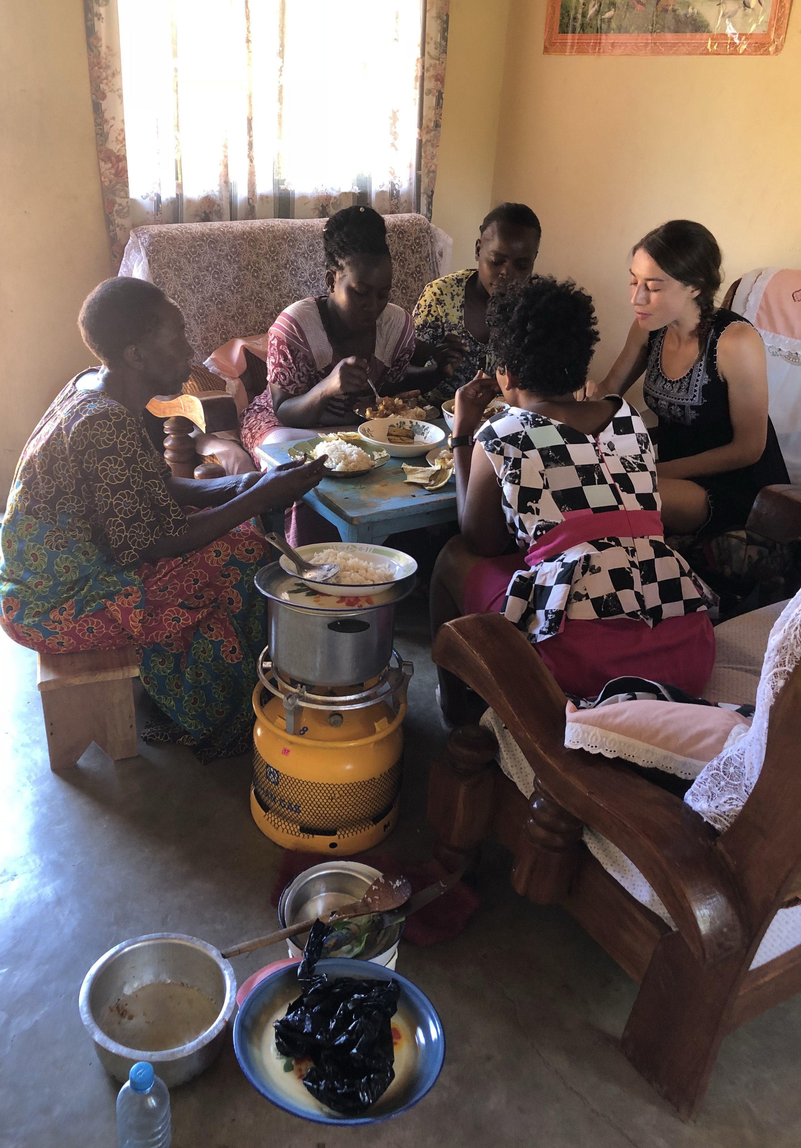 4 African women and a white woman eating around a table with a cookstove in foreground