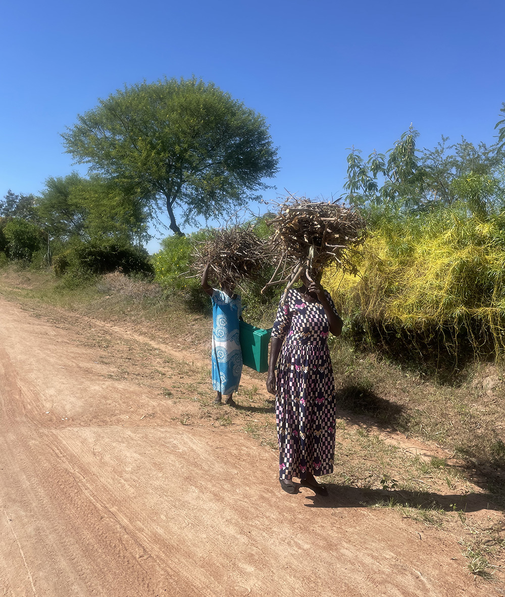 2 women walking alongside a red dirt road, carrying sticks on their heads