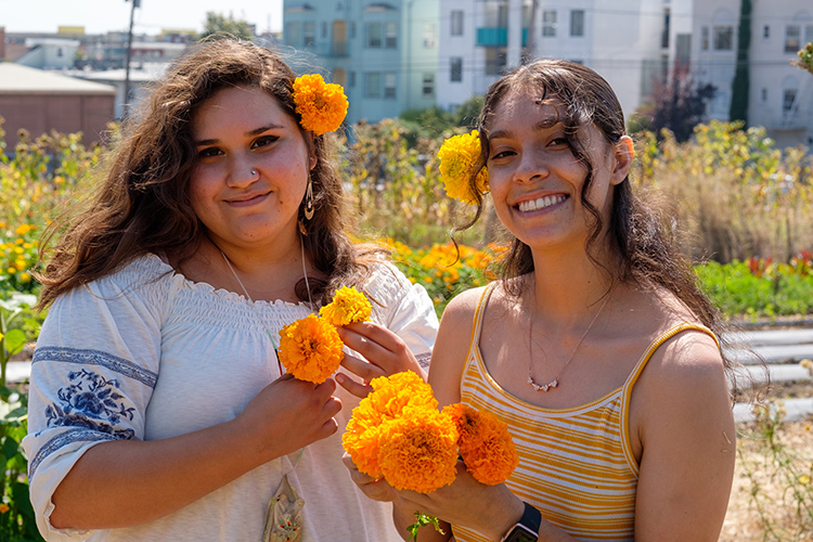 Two students holding marigolds and wearing marigolds in their hair pose for a photo in the Indigenous Community Learning Garden.