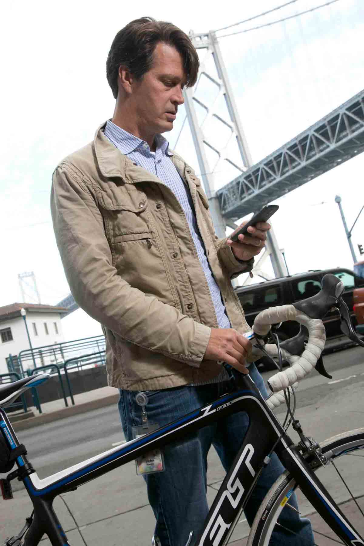 John Hanke standing next to a bike and holding an iPhone he is looking down at in front of a silver bridge.