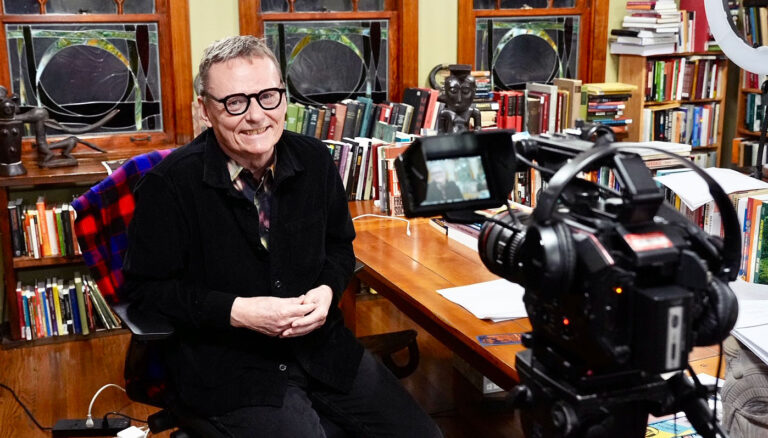 Economist James A. Robinson, sitted at a desk in a happily cluttered office, smiling as he waits to start a video interview