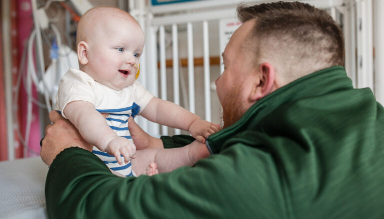 a man in a green hoodie holding a smiling baby with both arms