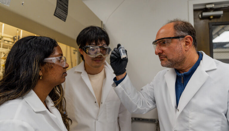 three people in white coats and goggles looking at a dark residue in a clear bottle