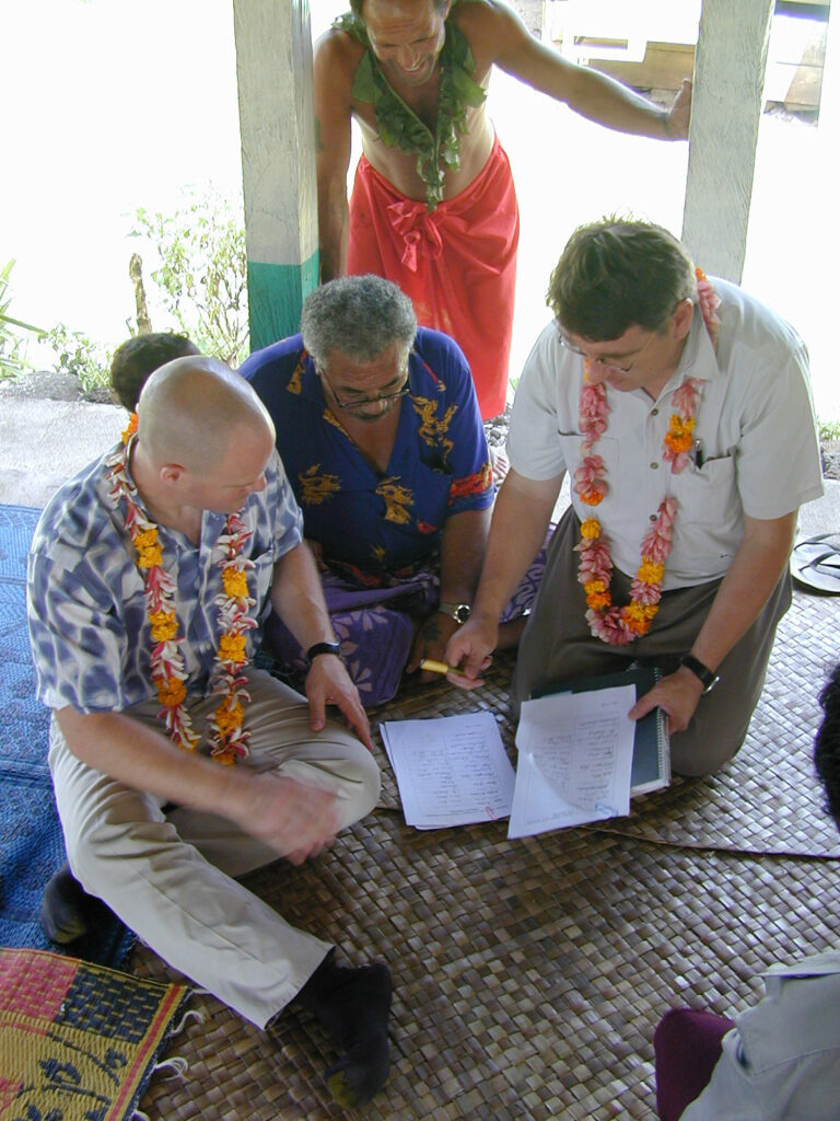 three men wearing leis kneeling and discussing papers on a mat, with a man standing behind them in a red skirt and green neck decoration