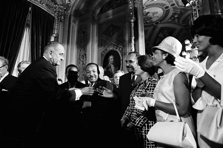 a black and white historical photo featuring President Lyndon Baines Johnson (left), the Rev. Dr. Martin Luther King (center) and other civil rights leaders in the ornate President's Room of the U.S. Capitol following Johnson's signing of the Voting Right