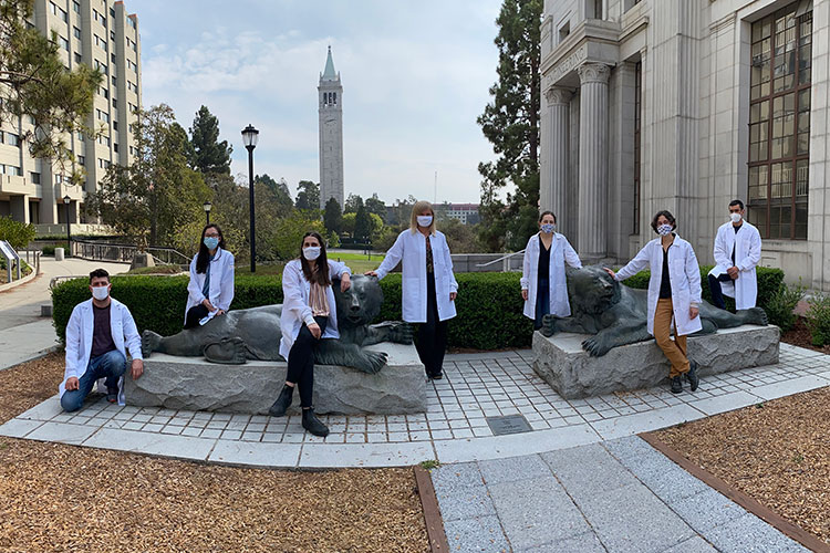 A photo of seven individuals wearing lab coats on the Berkeley campus. Each person is wearing a mask and they are all practicing physical distancing.