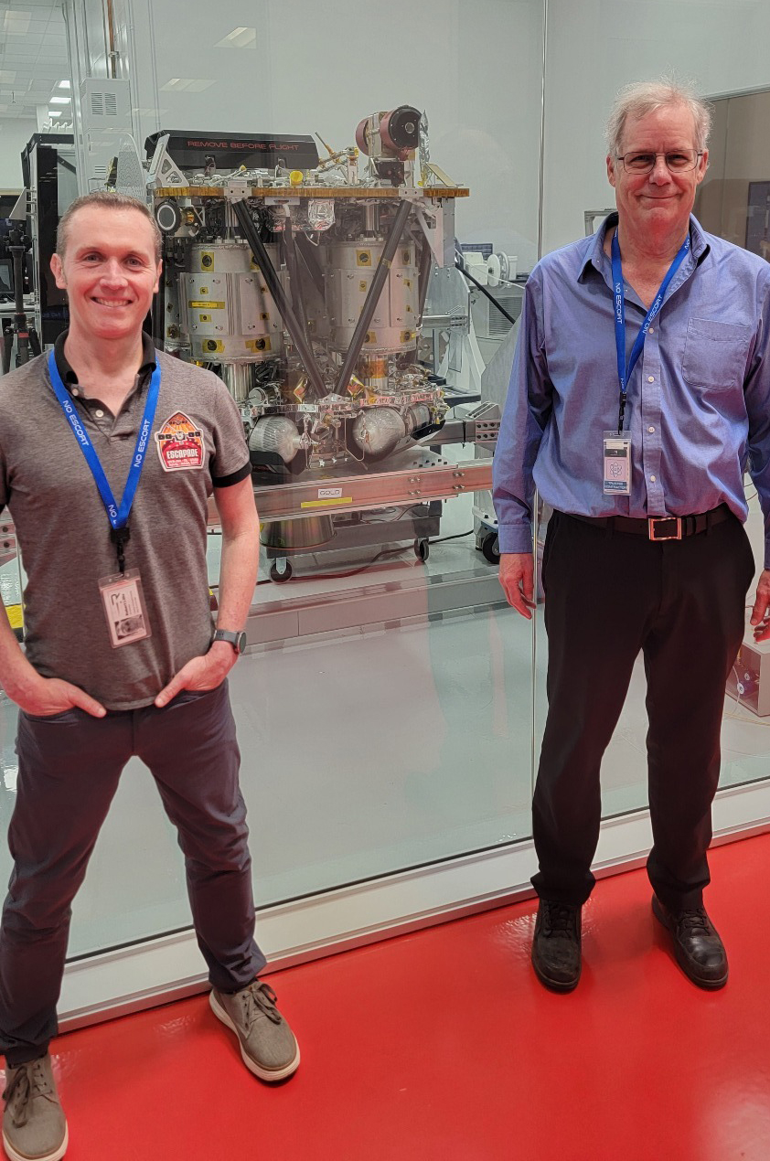 two men with name tags hanging around their necks pose in front of a piece of hardware