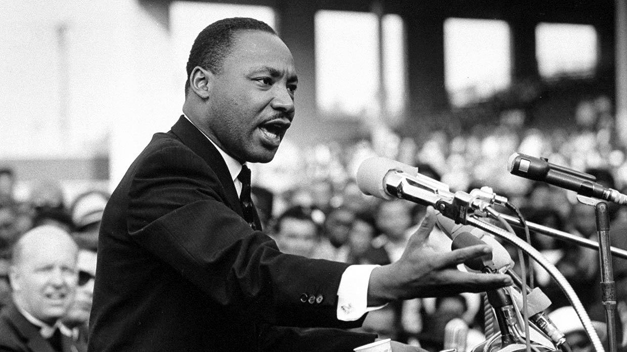 Martin Luther King stands before a bank of microphones and gestures passionately as he speaks to large crowd.