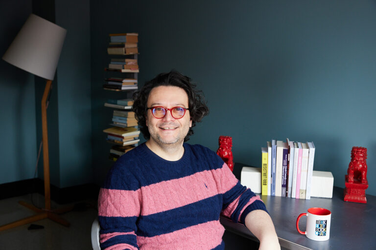 Mario Telò, professor of ancient Greek and Roman studies, rhetoric and comparative literature, sits at a desk wearing a pink and navy striped sweater and red frame eyeglasses. He is smiling and has books on his desk flanked by red Asian dragon statues.