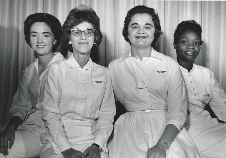 A black and white photo of three white woman and one Black woman, all with 1960s hair and wearing white uniforms.