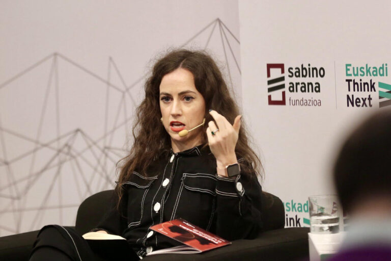 A brunette woman wearing a microphone speaks and gesticulates while speaking at a panel.