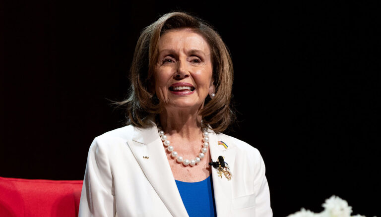 Former U.S. House Speaker Nancy Pelosi smiles as she sits on a red chair, wearing a blue blouse and a white blazer.