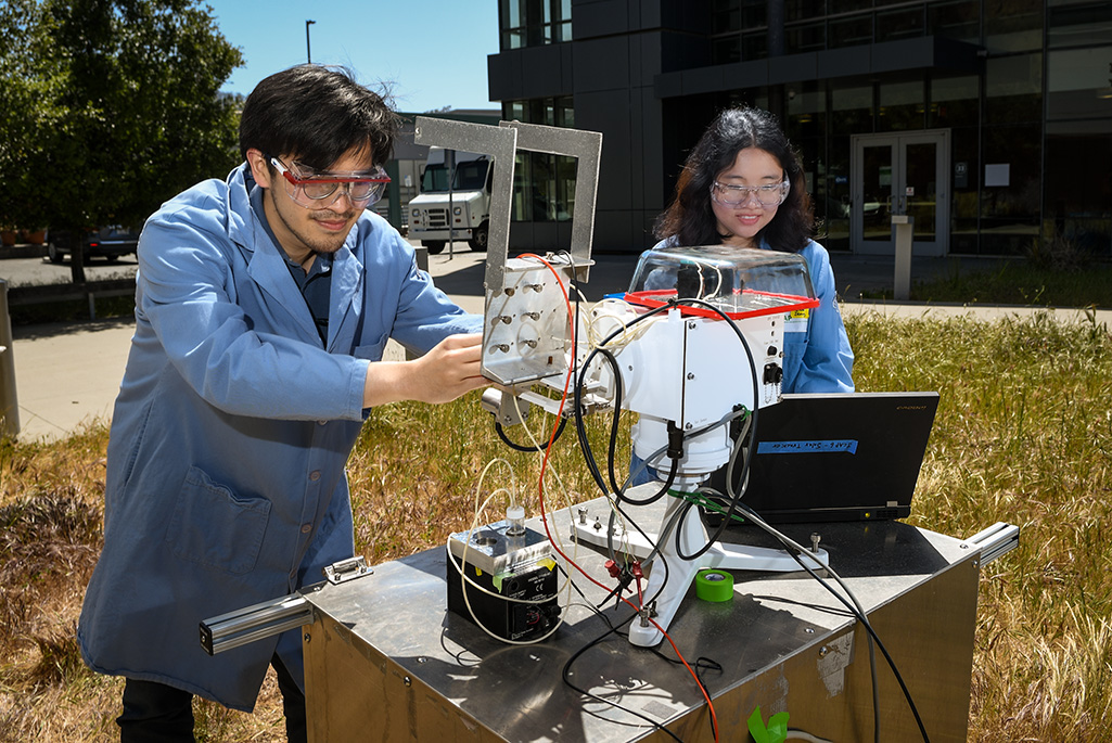 Two researchers in lab coats and goggles work with outdoor scientific equipment near a modern building.