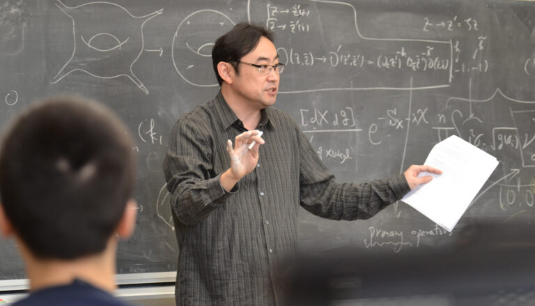 man at chalkboard gesturing to an audience with a piece of chalk