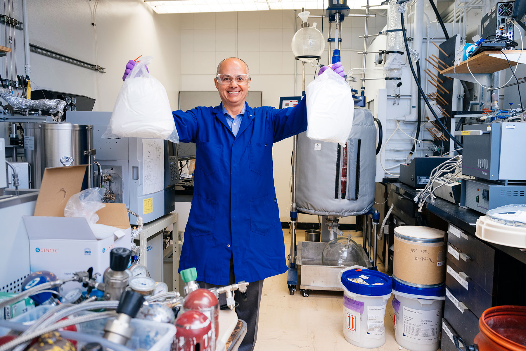 A person wearing a blue lab coat and safety glasses holds up two bags of white material and smiles.