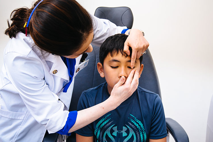 A photo shows a child sitting in an exam chair. An optometrist stands over the child, examining their eye.